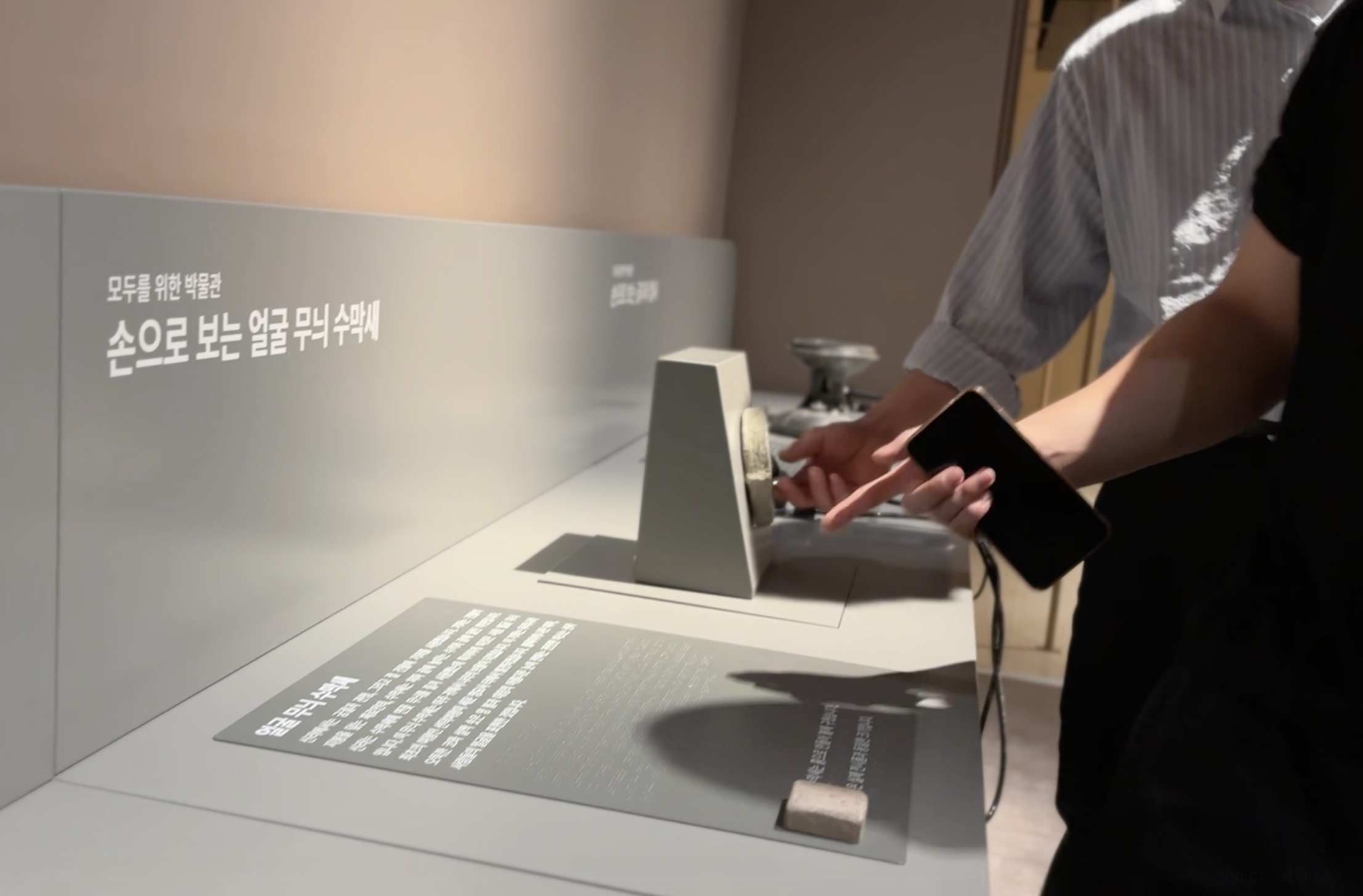 Foto horizontal de una exhibición de réplicas de piezas antiguas de Corea. Se ven las manos de 2 personas tocando una figura circular montada en un pequeño pedestal. A sus lados se aprecian otras piezas similares, y muestras de materiales rugosos junto a cédulas en braille y bocinas parecidas a las de un teléfono antiguo.
